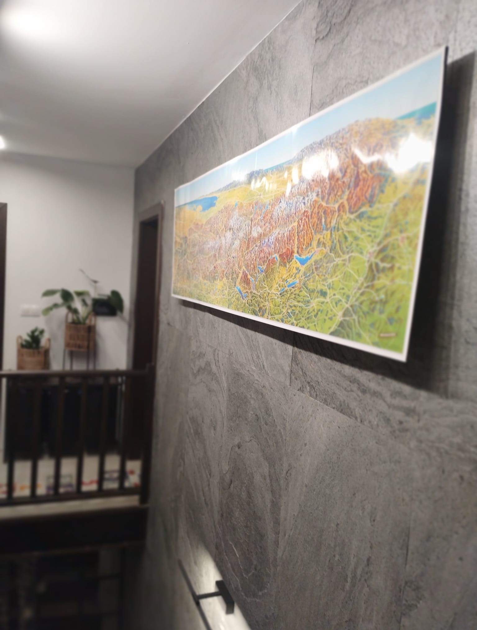 Długi, panoramiczny obraz z mapą krajobrazową wisi na szarej, teksturowanej ścianie. W tle widoczne schody i rośliny doniczkowe, tworząc nowoczesne wnętrze.