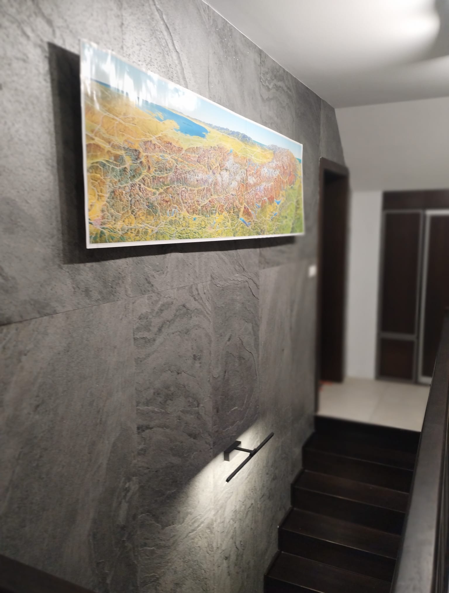 Szara ściana z kamienną okładziną, na której wisi podłużny obraz z krajobrazem. Widoczne drewniane schody i fragment poręczy.