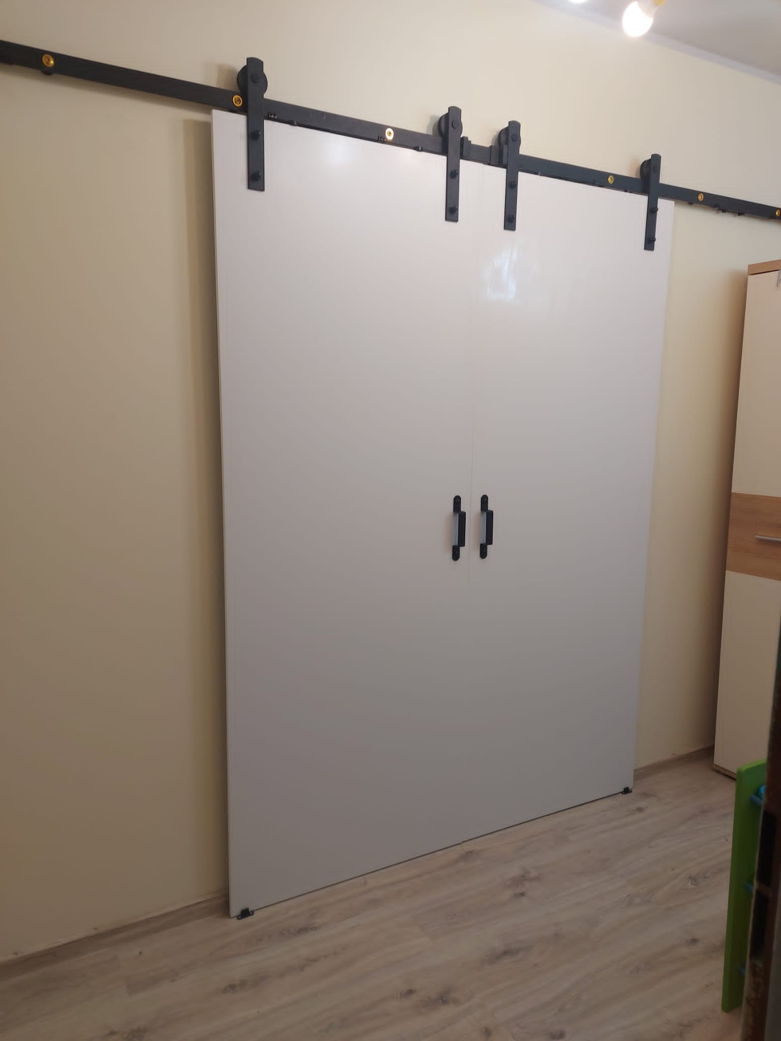 Białe, przesuwne drzwi na czarnej, metalowej szynie z uchwytami, zamontowane w beżowym wnętrzu. Widoczna podłoga i fragment szafy.