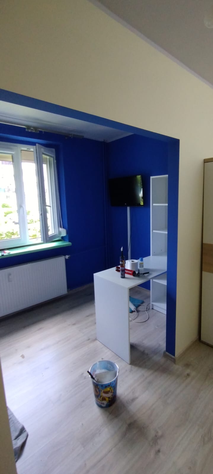 Pokój po malowaniu: ściany w odcieniach niebieskiego i beżu, białe meble, widoczny kubeł z farbą. Ujęcie z perspektywy podłogi, podkreślające świeżość i kontrast kolorów.