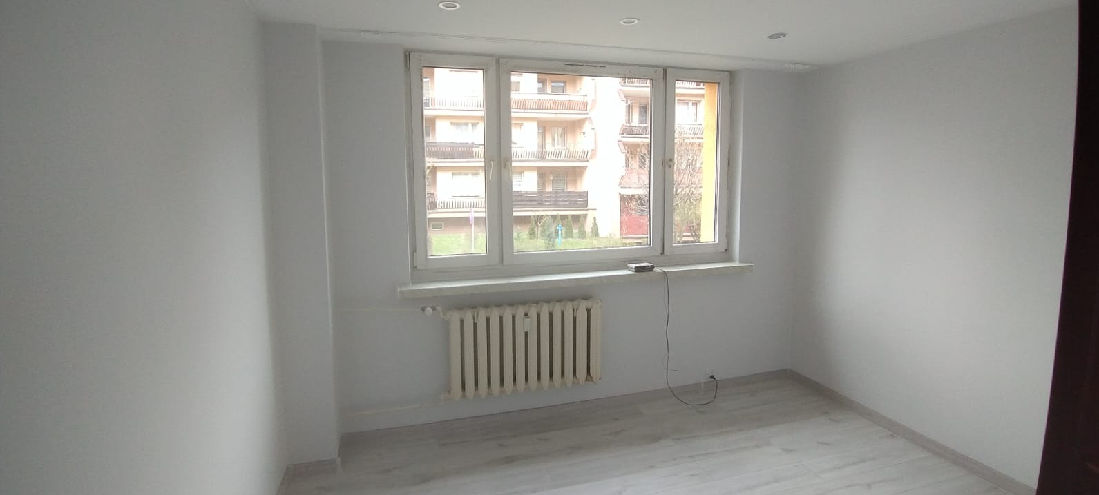 Pomalowane na biało puste pomieszczenie z grzejnikiem pod oknem. Świeżo wykończone ściany i jasna podłoga. Widok na budynek zza okna.