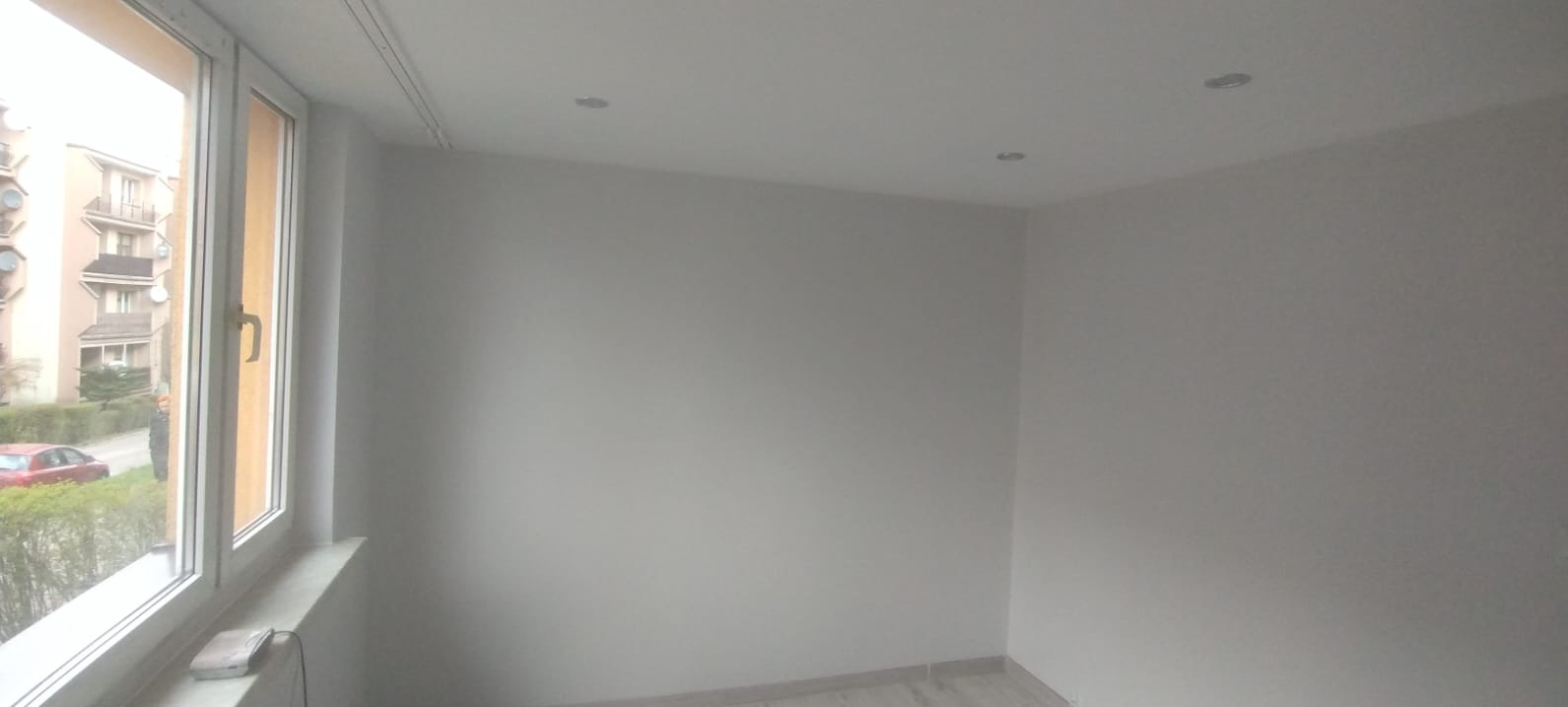 Jasne wnętrze po malowaniu: białe ściany, okno z widokiem na budynek mieszkalny i zaparkowany czerwony samochód, w suficie zamontowane trzy halogeny.