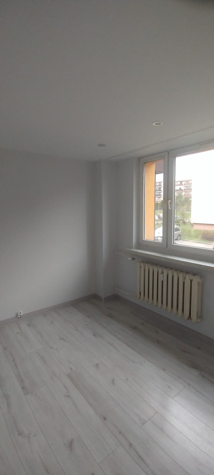 Jasne, świeżo wyremontowane wnętrze z panelami podłogowymi, białymi ścianami i grzejnikiem pod oknem. Widok na zewnątrz przez okno.