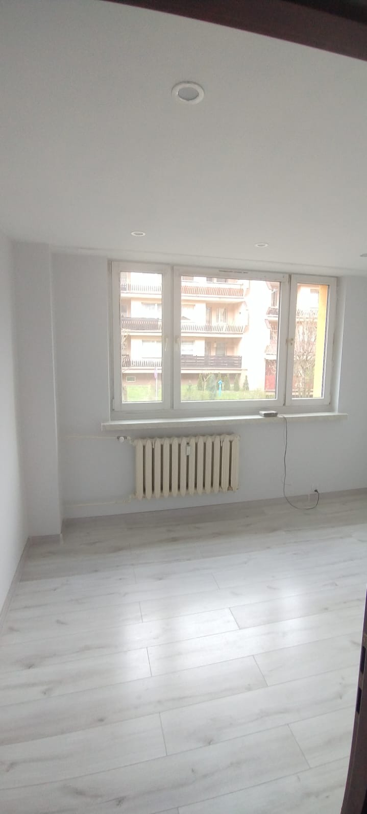 Jasne, odnowione wnętrze pokoju z białymi ścianami, panelami podłogowymi i grzejnikiem pod oknem. Widok na budynek zza okna.