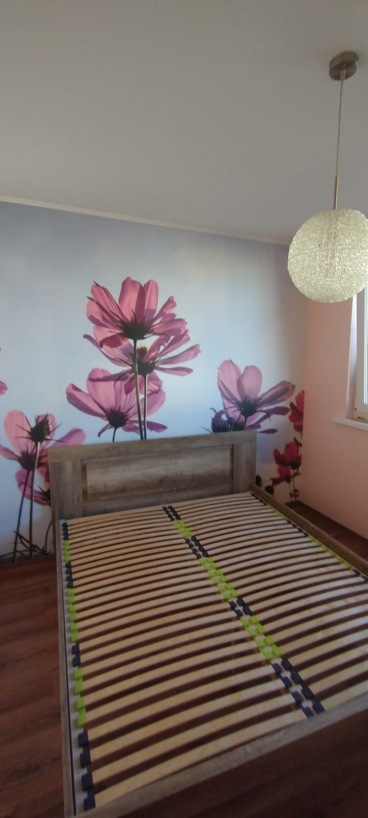 Jasne drewniane łóżko bez materaca, widok na ścianę z fototapetą w kwiaty i okrągłą lampę. Minimalistyczny wystrój sypialni w pastelowych barwach.