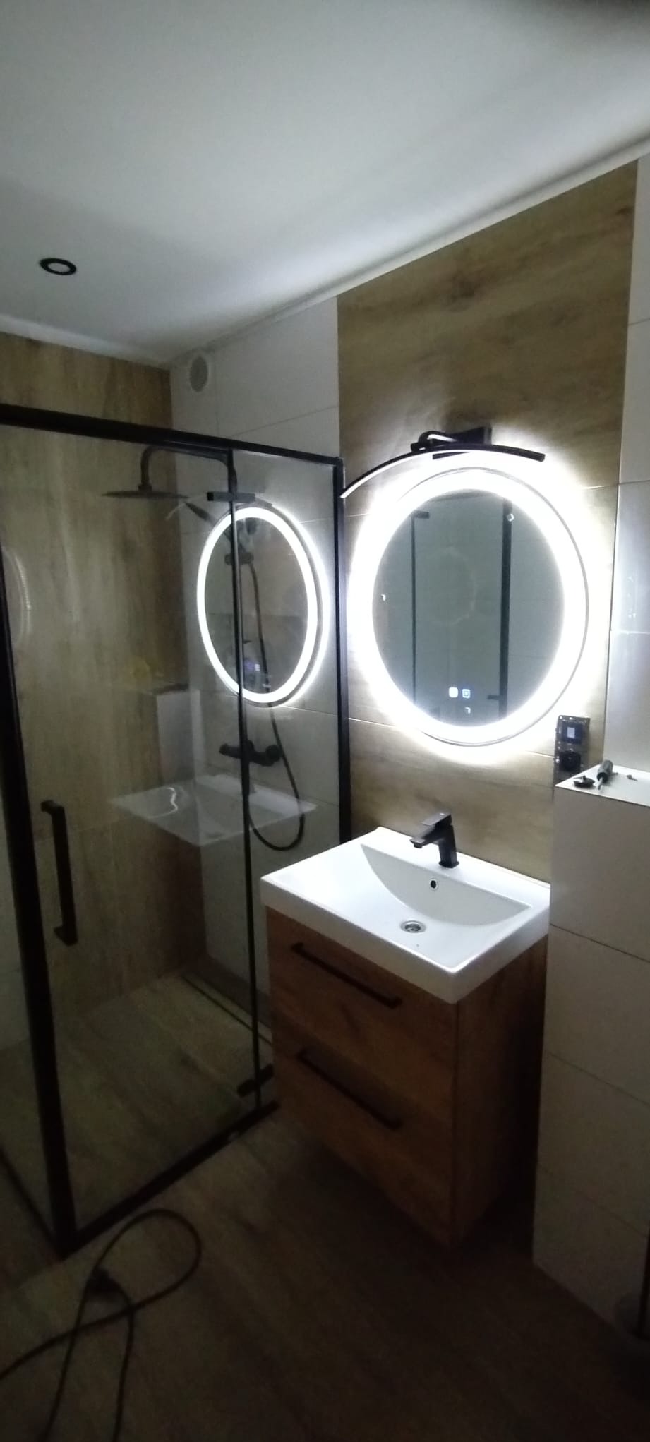 Nowoczesna łazienka z kabiną prysznicową typu walk-in, umywalką na drewnianej szafce i okrągłymi lustrami z podświetleniem LED, płytki imitujące drewno na ścianach.