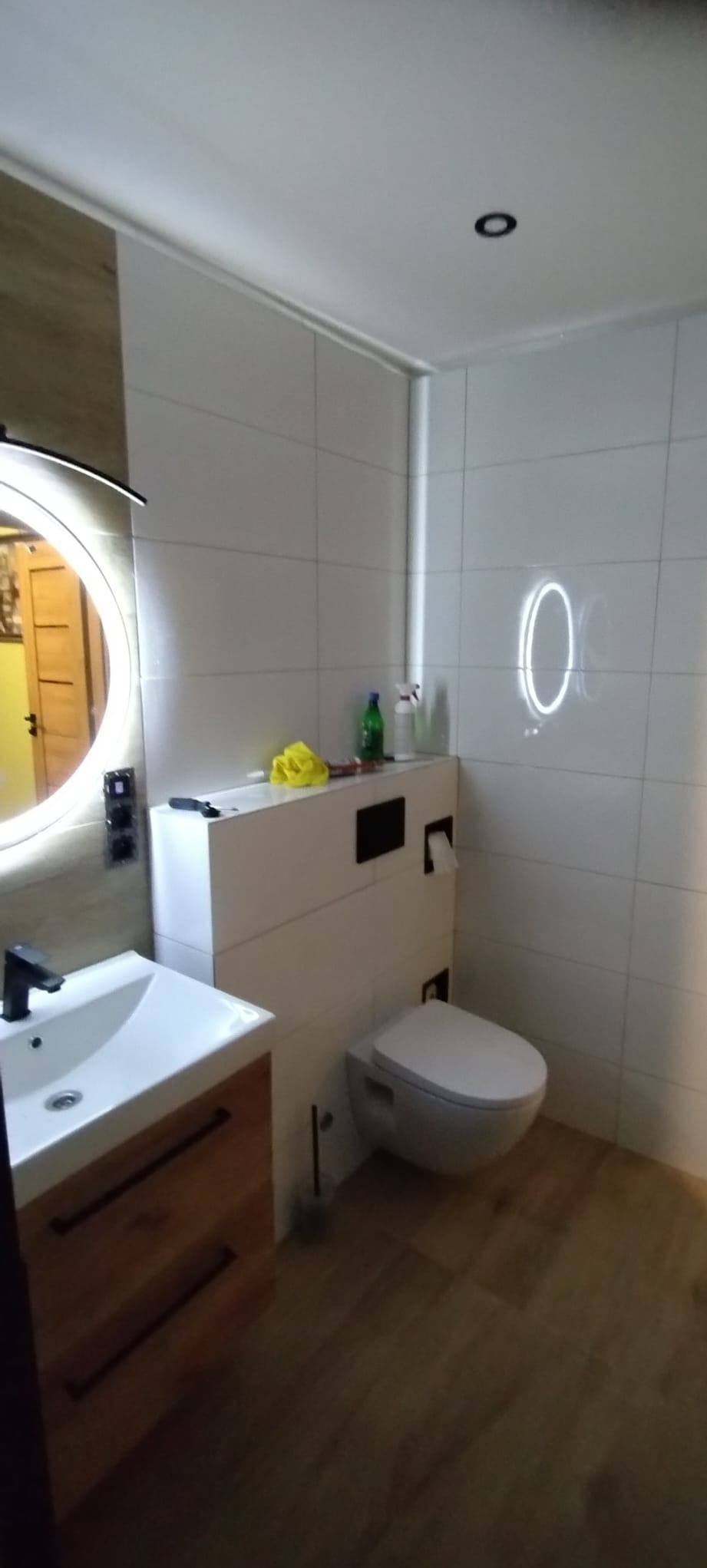 Nowoczesna łazienka z okrągłym lustrem LED, drewnianą szafką pod umywalką i podwieszaną toaletą. Białe płytki na ścianach i drewnopodobna podłoga.