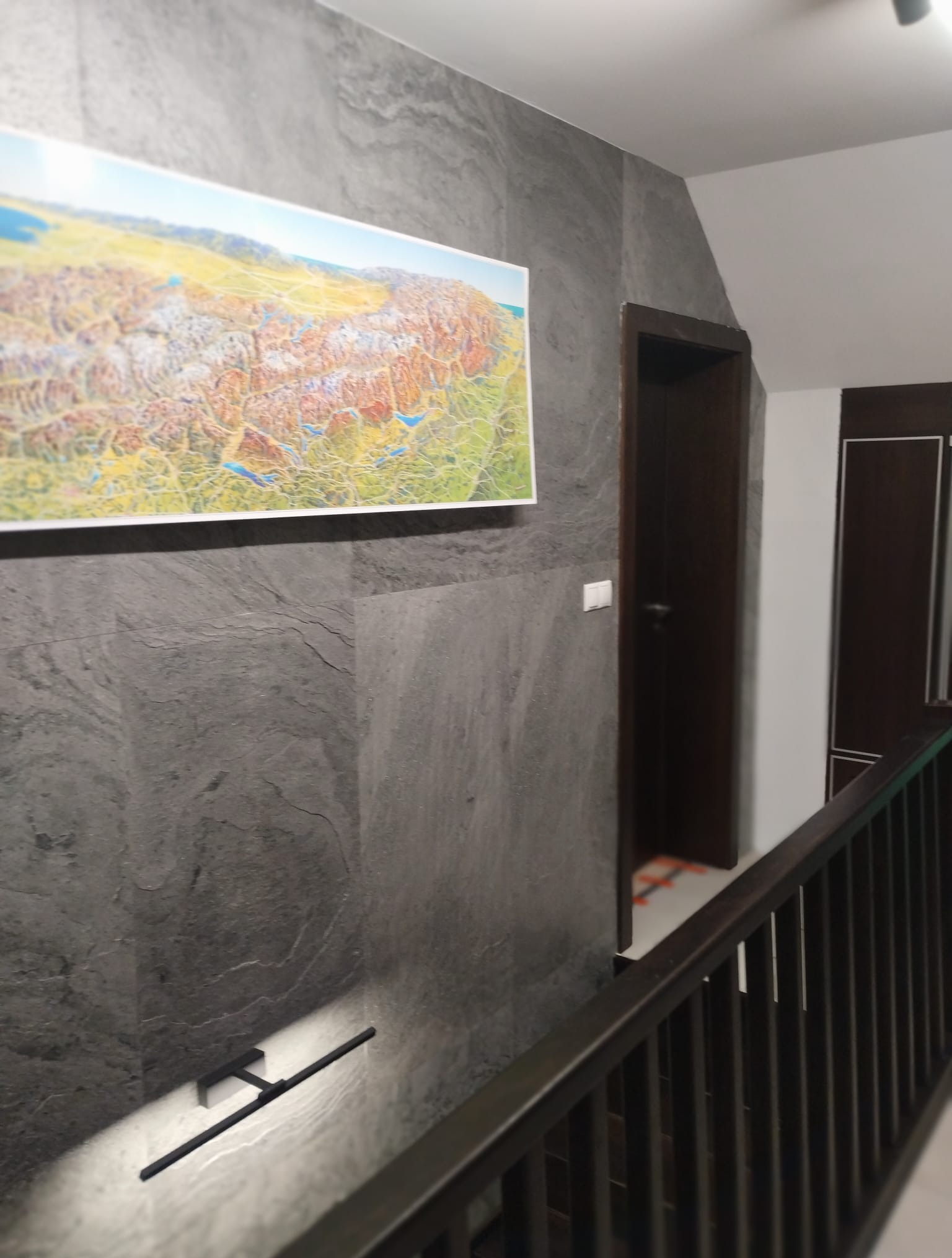 Ściana wykończona szarym kamieniem dekoracyjnym z podświetleniem, na której wisi obraz. W tle widoczne drzwi i fragment balustrady schodów.