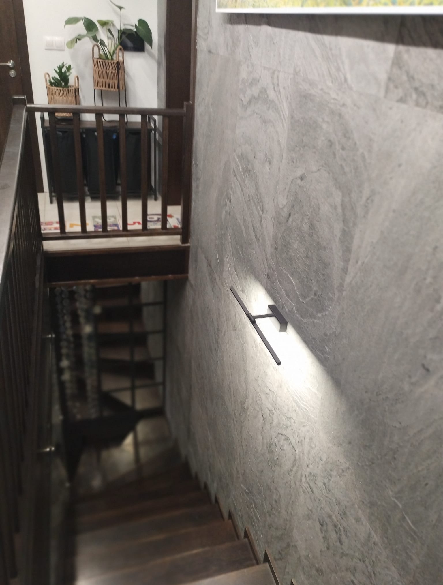 Klatka schodowa z drewnianymi schodami i balustradą. Ściana wykończona szarą płytą kamienną z oświetleniem punktowym. W tle widoczne rośliny doniczkowe.