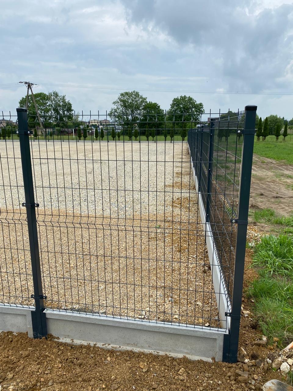 Ciemnoszare ogrodzenie panelowe na betonowej podmurówce, widok z boku. W tle drzewa i pochmurne niebo. Teren wysypany żwirem.