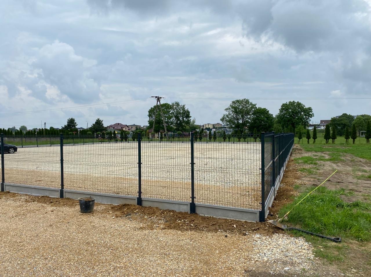 Nowoczesne ogrodzenie panelowe z podmurówką na żwirowym podłożu. Widoczne słupki i siatka. W tle drzewa i zabudowania mieszkalne.