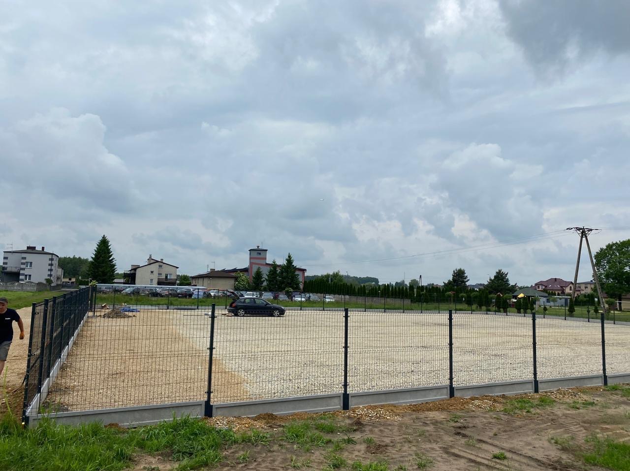 Nowe ogrodzenie panelowe z podmurówką betonową otacza teren wysypany żwirem, z budynkami w tle i pochmurnym niebem.
