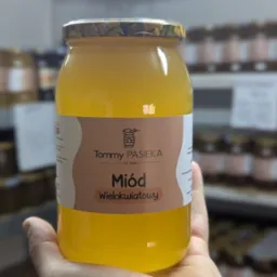 Słoik wielokwiatowego miodu od Tommy Pasieka, trzymany w dłoni, z półkami pełnymi słoików w tle. Widoczna etykieta i złocista barwa miodu.
