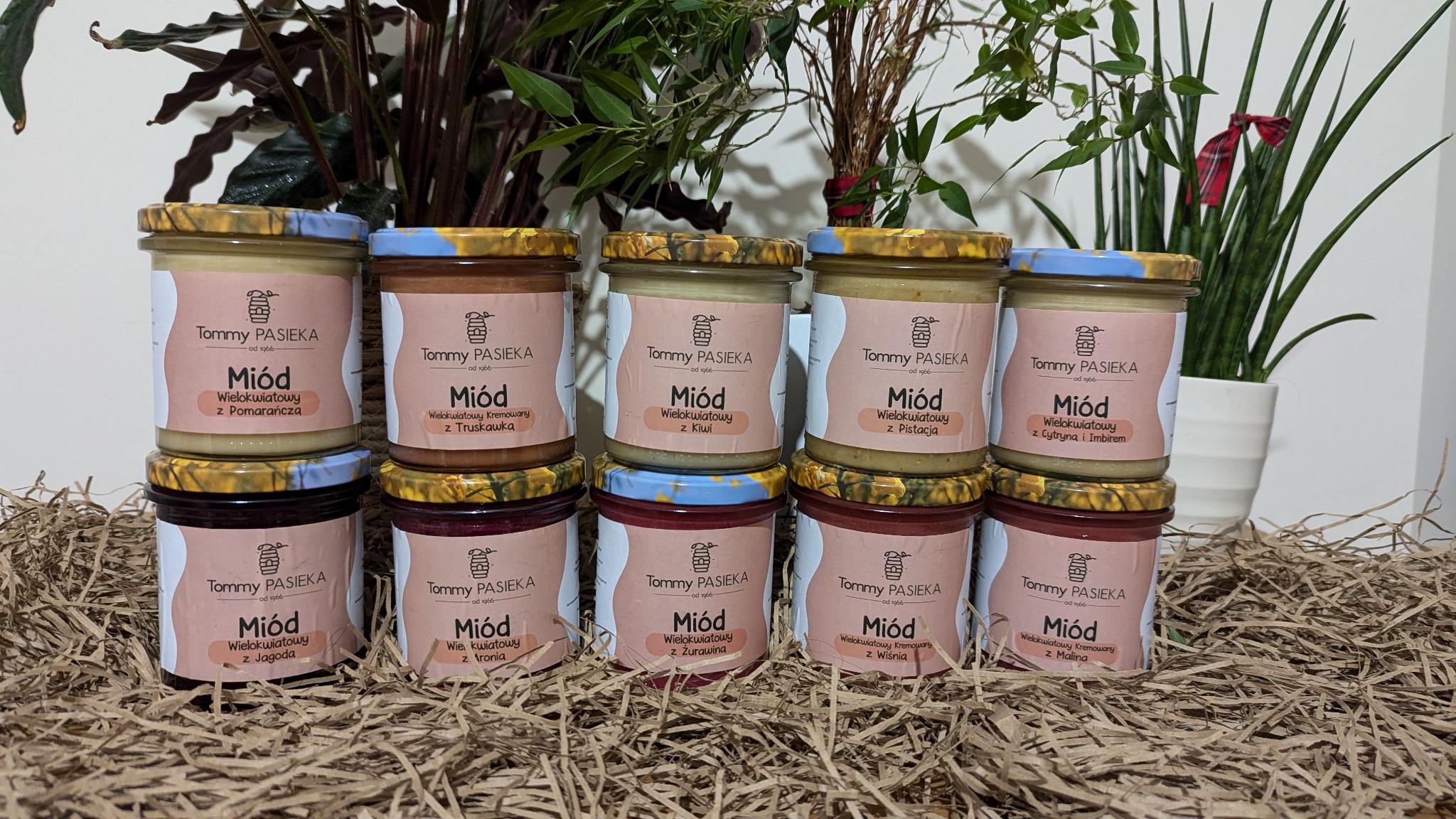Zestaw miodów wielokwiatowych w słoikach, różne smaki, etykiety z nazwą pasieki, tło z roślin i tektury falistej.