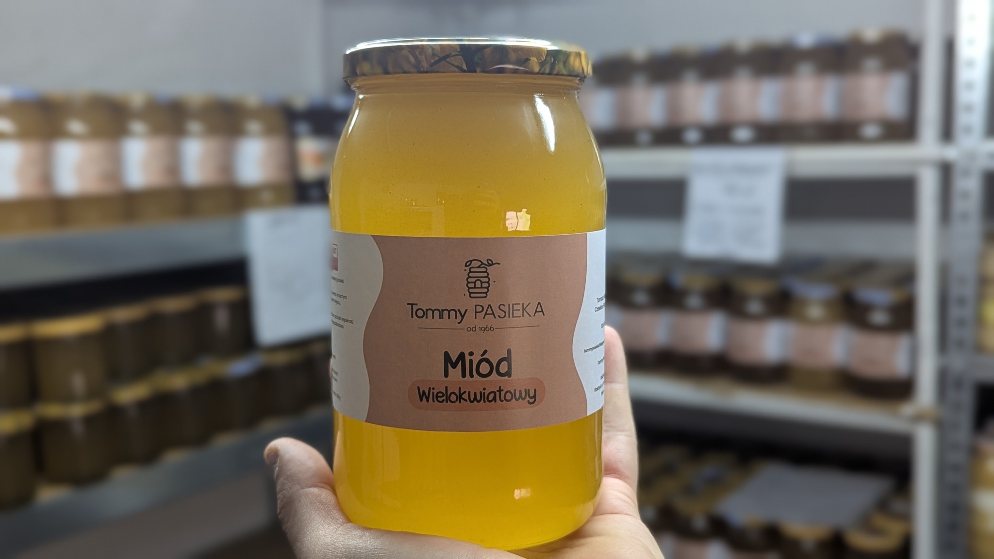 Słoik wielokwiatowego miodu od Tommy Pasieka, trzymany w dłoni, z półkami pełnymi słoików w tle. Widoczna etykieta i złocista barwa miodu.