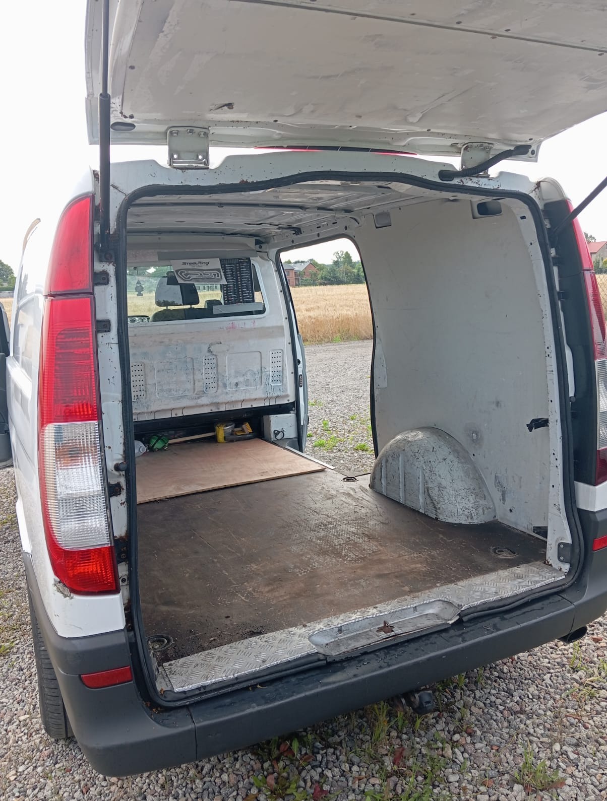 Otwarta przestrzeń ładunkowa białego busa, z widoczną podłogą i narzędziami. W tle pole uprawne i budynki. Widok z tyłu pojazdu.