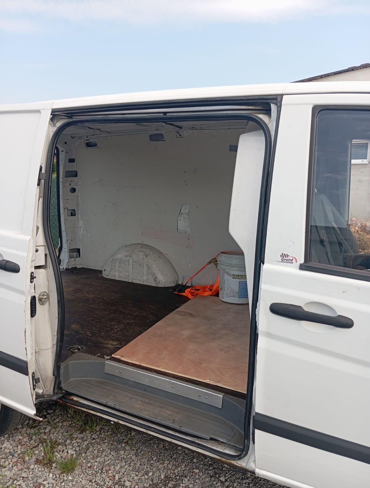 Otwarty, biały furgon z pustą przestrzenią ładunkową, przygotowany do załadunku. Widoczne zabezpieczenia ładunku i częściowo widoczne tło z budynkiem.
