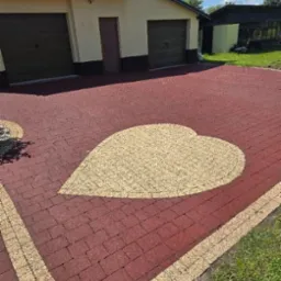 Czerwona kostka brukowa z beżowym sercem na podjeździe do garażu. Ujęcie z poziomu gruntu, widoczny fragment budynku.
