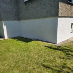 Ocieplony i otynkowany fragment elewacji budynku z boiskiem do koszykówki, widoczne z poziomu trawnika. Jasna, świeża elewacja kontrastuje z szarą, starszą górną częścią.