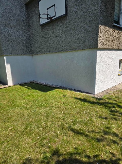 Ocieplony i otynkowany fragment elewacji budynku z boiskiem do koszykówki, widoczne z poziomu trawnika. Jasna, świeża elewacja kontrastuje z szarą, starszą górną częścią.