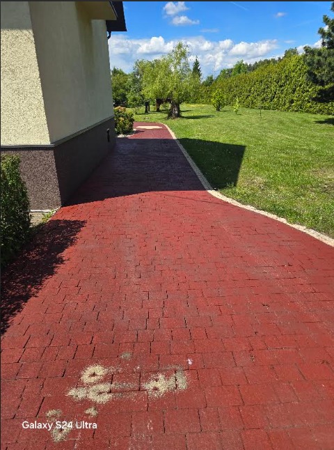 Czerwony chodnik z kostki brukowej prowadzi wzdłuż jasnego budynku z ciemnym cokołem, w tle zielony trawnik i drzewa. Fragment chodnika pokryty piaskiem.