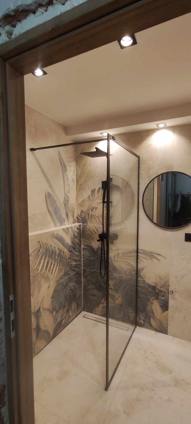 Nowoczesna kabina prysznicowa z czarnymi detalami i tropikalnym motywem na płytkach. Okrągłe lustro dopełnia aranżację łazienki.