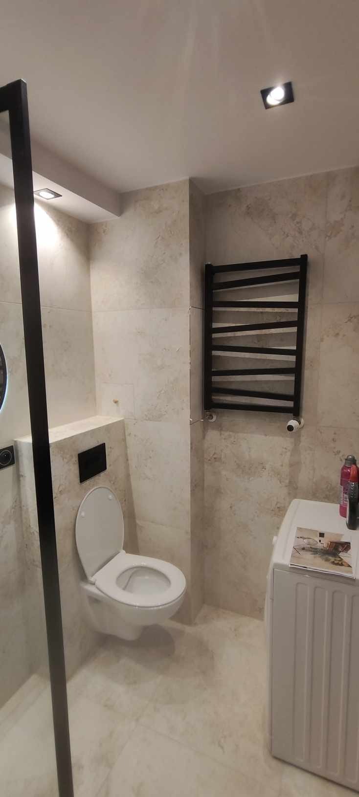 Elegancka łazienka z beżowymi płytkami, podwieszaną toaletą i czarnym grzejnikiem drabinkowym. Kabina prysznicowa z czarną ramą. Nowoczesny design.