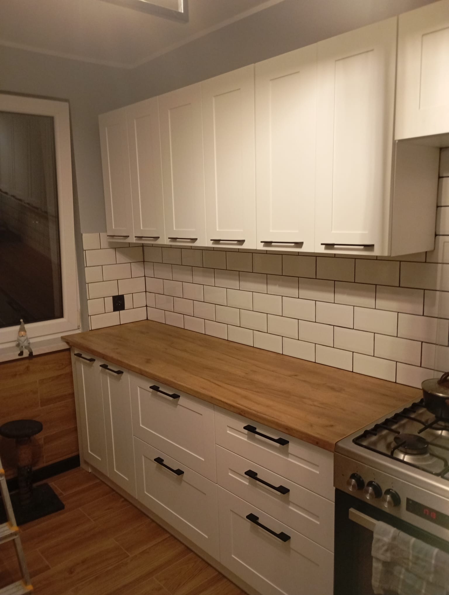 Jasna kuchnia z drewnianym blatem i białymi szafkami z czarnymi uchwytami. Na ścianie białe płytki imitujące cegłę. Widoczna kuchenka gazowa i fragment podłogi.