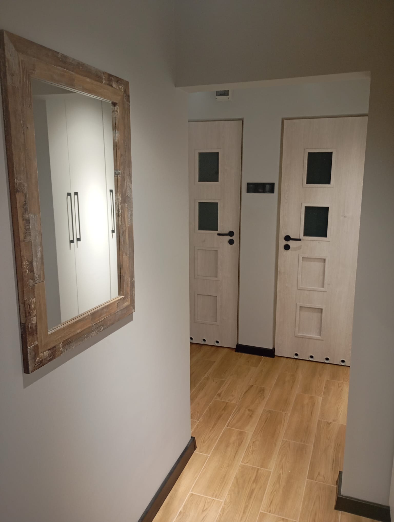 Korytarz po remoncie: drewniana podłoga, dwoje drzwi z czarnymi klamkami i lustro w drewnianej ramie na ścianie. Widok z perspektywy.