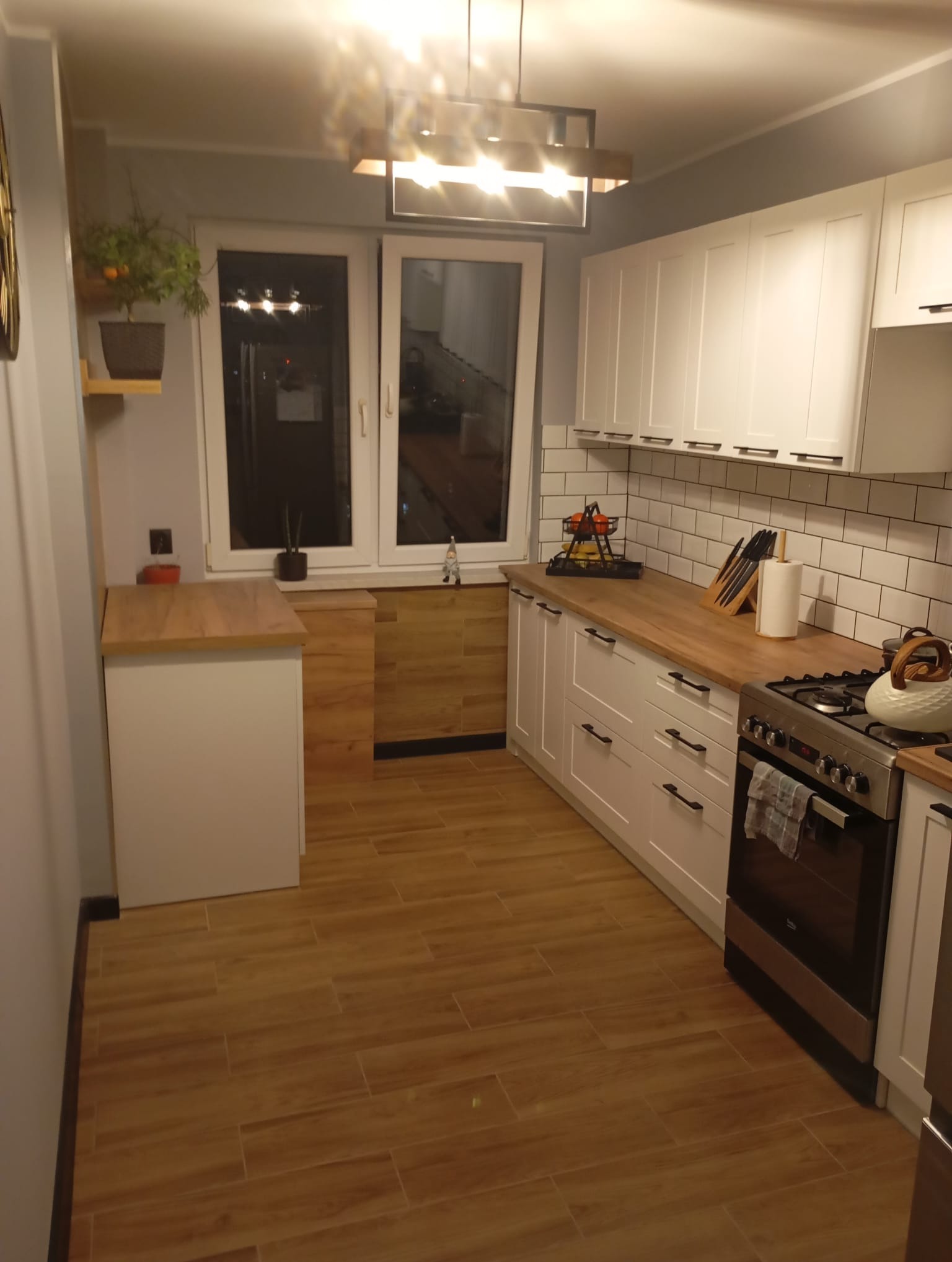 Jasna kuchnia po remoncie: białe szafki z drewnianym blatem, płytki metro na ścianie, drewnopodobna podłoga. Widok na okno z roślinami.