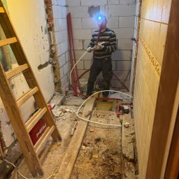 Dom-mur usługi ogólnobudowlane Krzysztof Snoch - Instalator z czołówką montuje rury w surowym wnętrzu łazienki z widoczną drabiną i fragmentami ścian z pustaków oraz płytek. Etap prac instalacyjnych.
