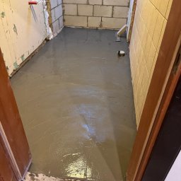 Dom-mur usługi ogólnobudowlane Krzysztof Snoch - Wylany, świeży podkład podłogowy w pomieszczeniu z fragmentami ścian z bloczków i płytek. Widoczne narzędzia do rozprowadzania wylewki.