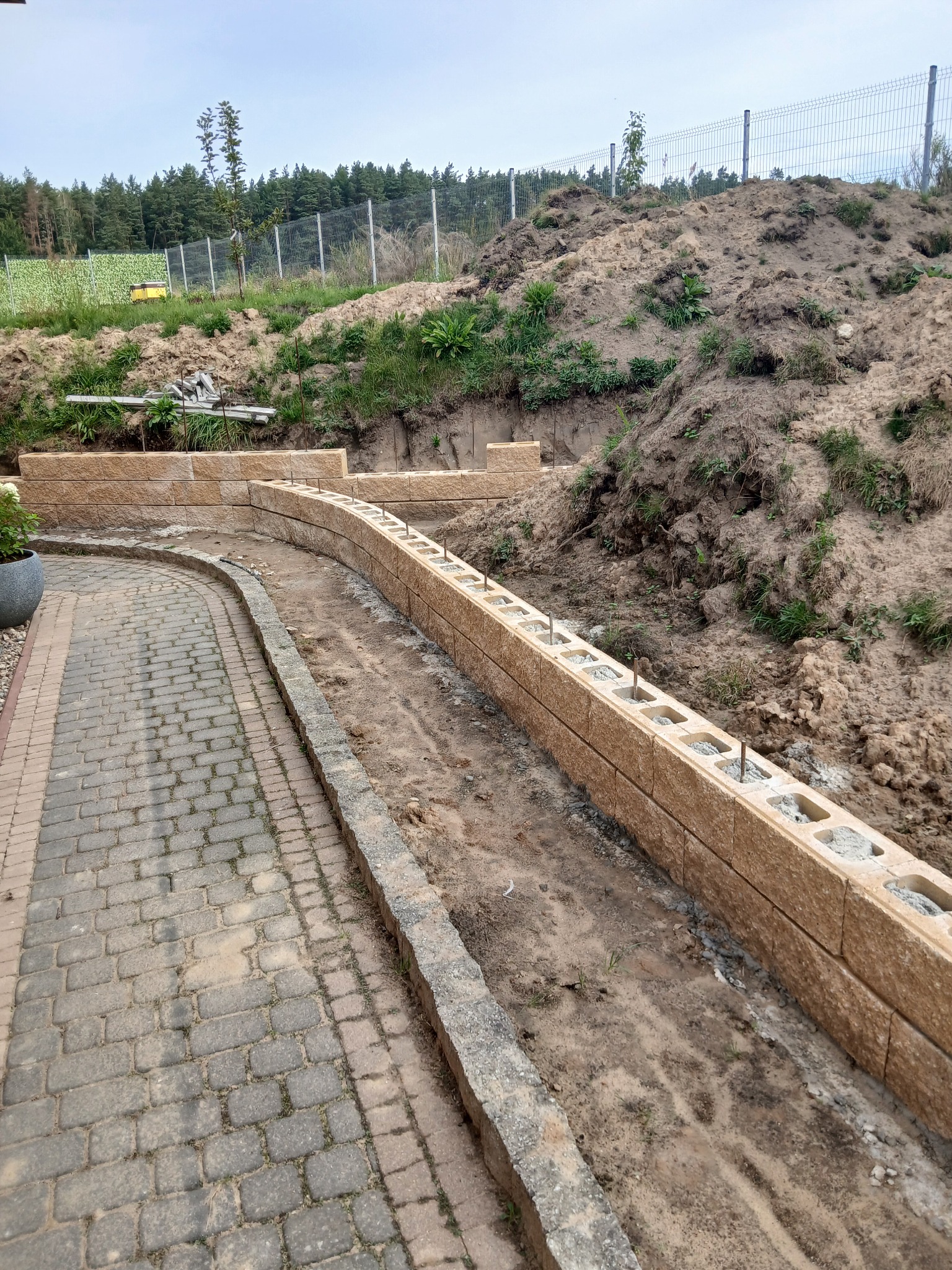 Kaskadowy murek oporowy z pustaków betonowych, częściowo wypełnionych, z wystającymi prętami zbrojeniowymi, obok ścieżki z kostki brukowej i skarpy ziemnej.