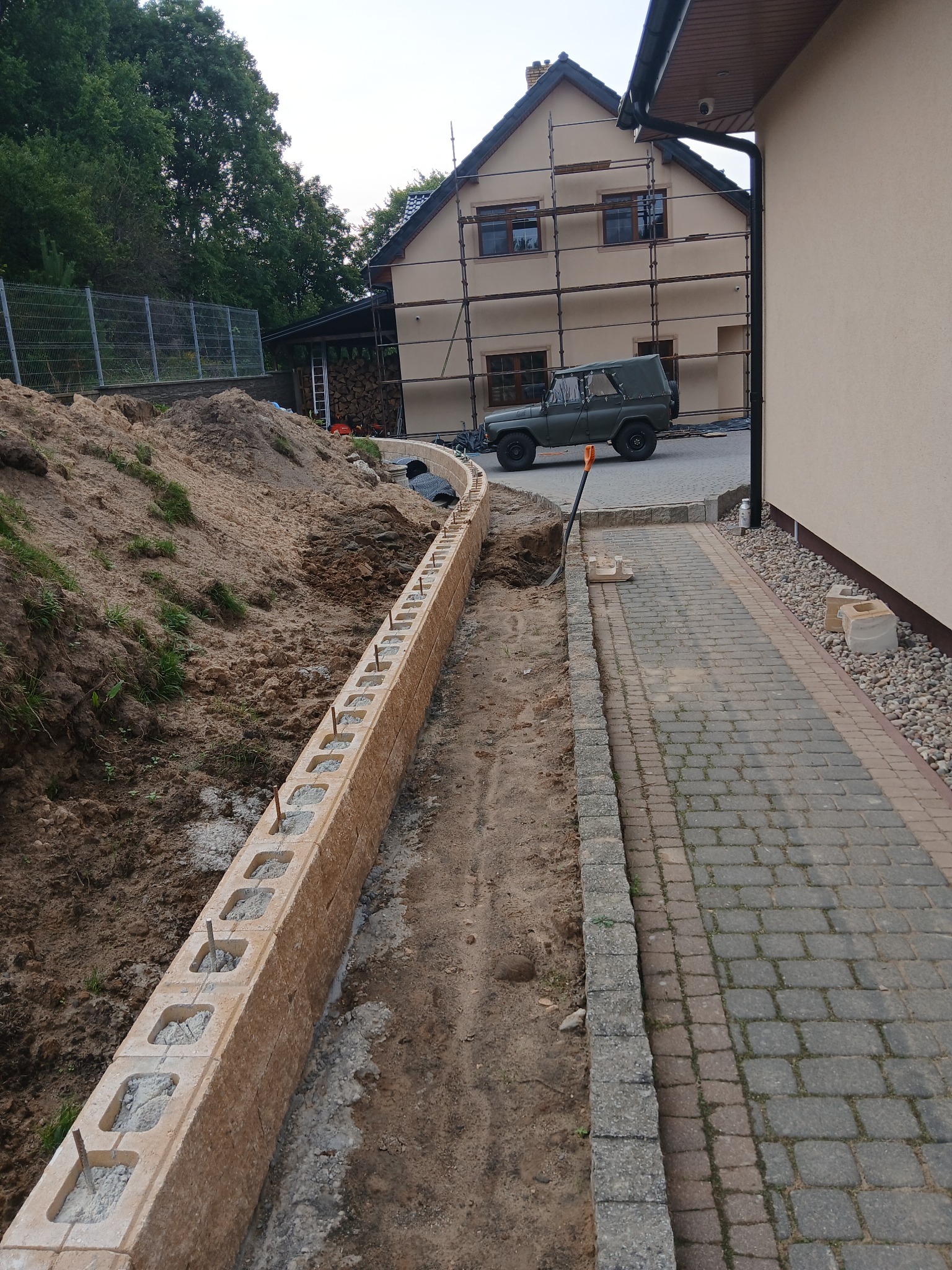 Wykop pod fundament z częściowo ułożonymi bloczkami betonowymi z wystającymi prętami zbrojeniowymi. W tle dom w trakcie budowy ze stojącym samochodem.