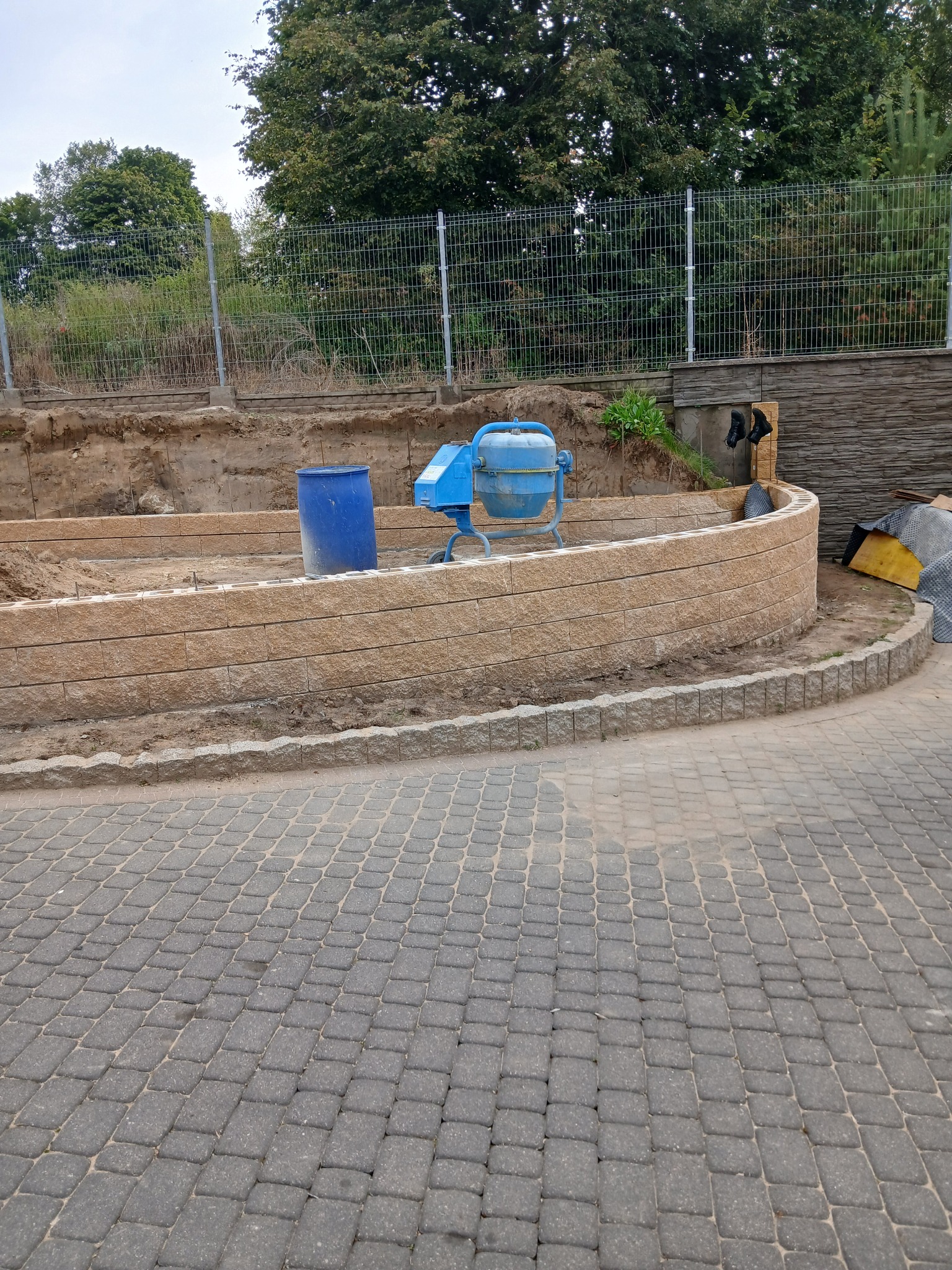 Budowa murku oporowego z kostki, widoczna betoniarka i kostka brukowa. Etap prac ziemnych, w tle ogrodzenie panelowe i drzewa. Ujęcie z góry.