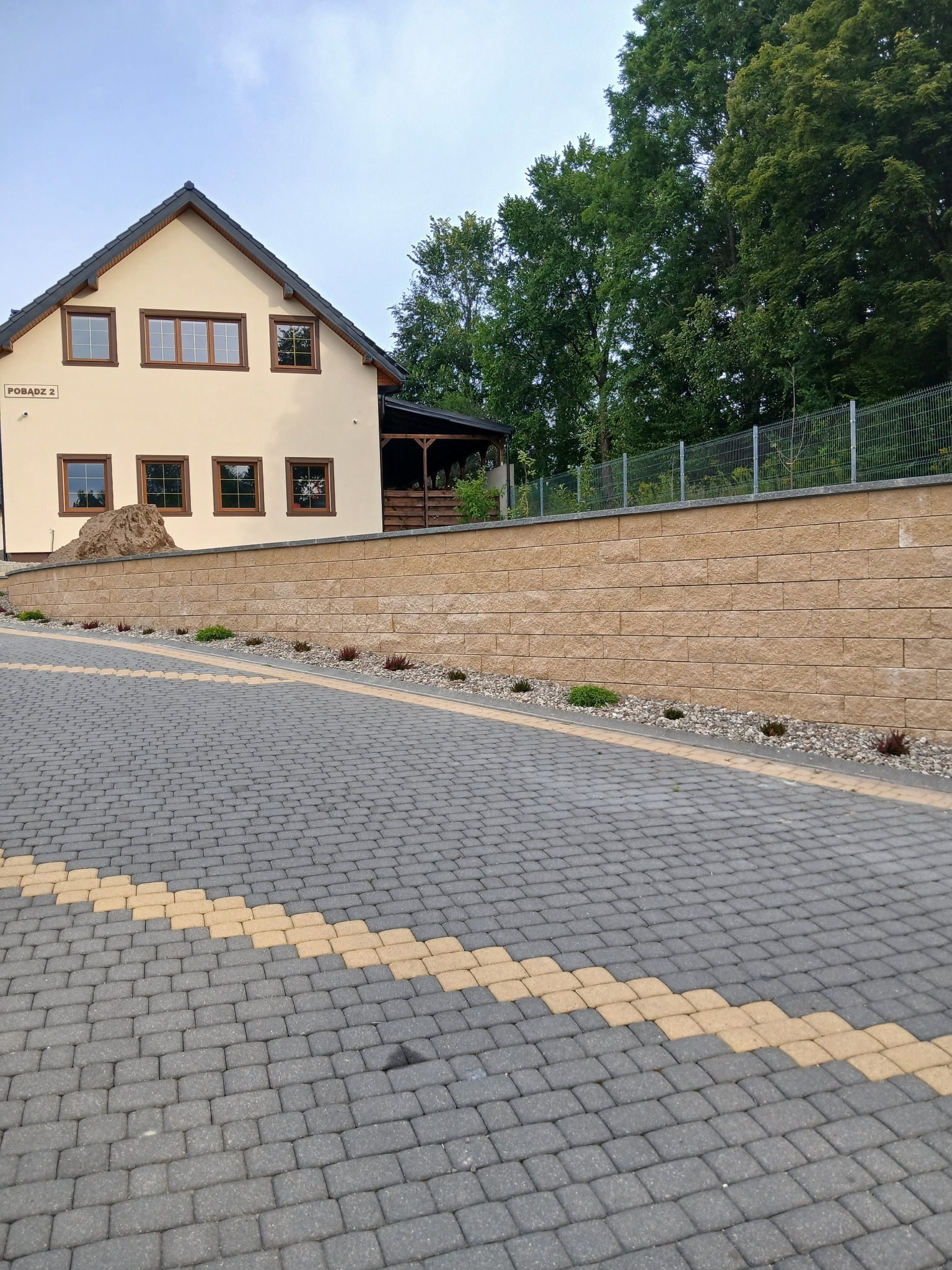 Ukończona posesja z szarej kostki brukowej z żółtymi akcentami, mur oporowy z jasnego kamienia i dom z beżową elewacją. Widok na posesję z dołu.