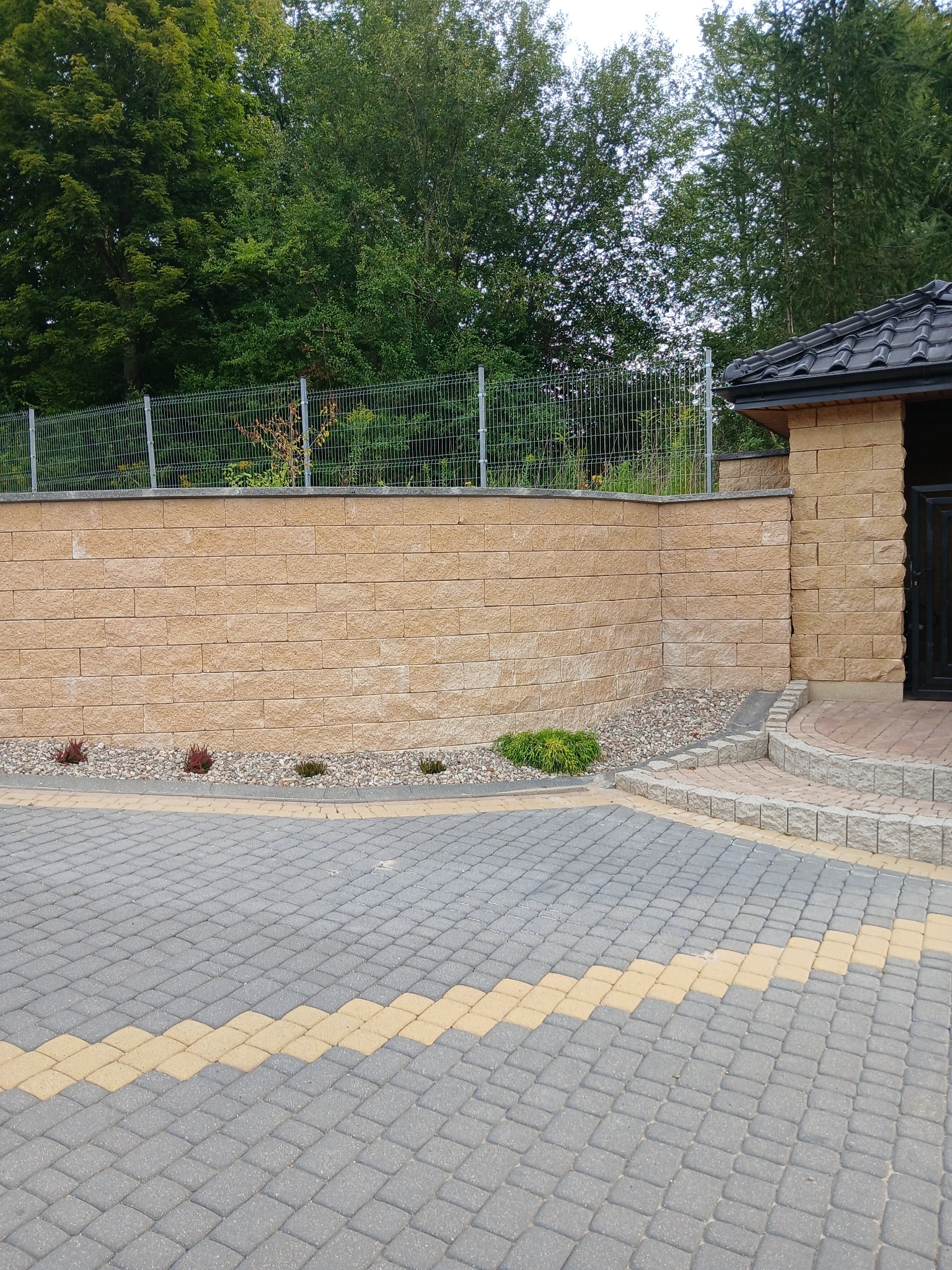 Ogród z murem oporowym z piaskowca, ogrodzeniem panelowym i ścieżką z kostki brukowej w odcieniach szarości i żółci. Uroku dodają małe krzewy i kamyczki.