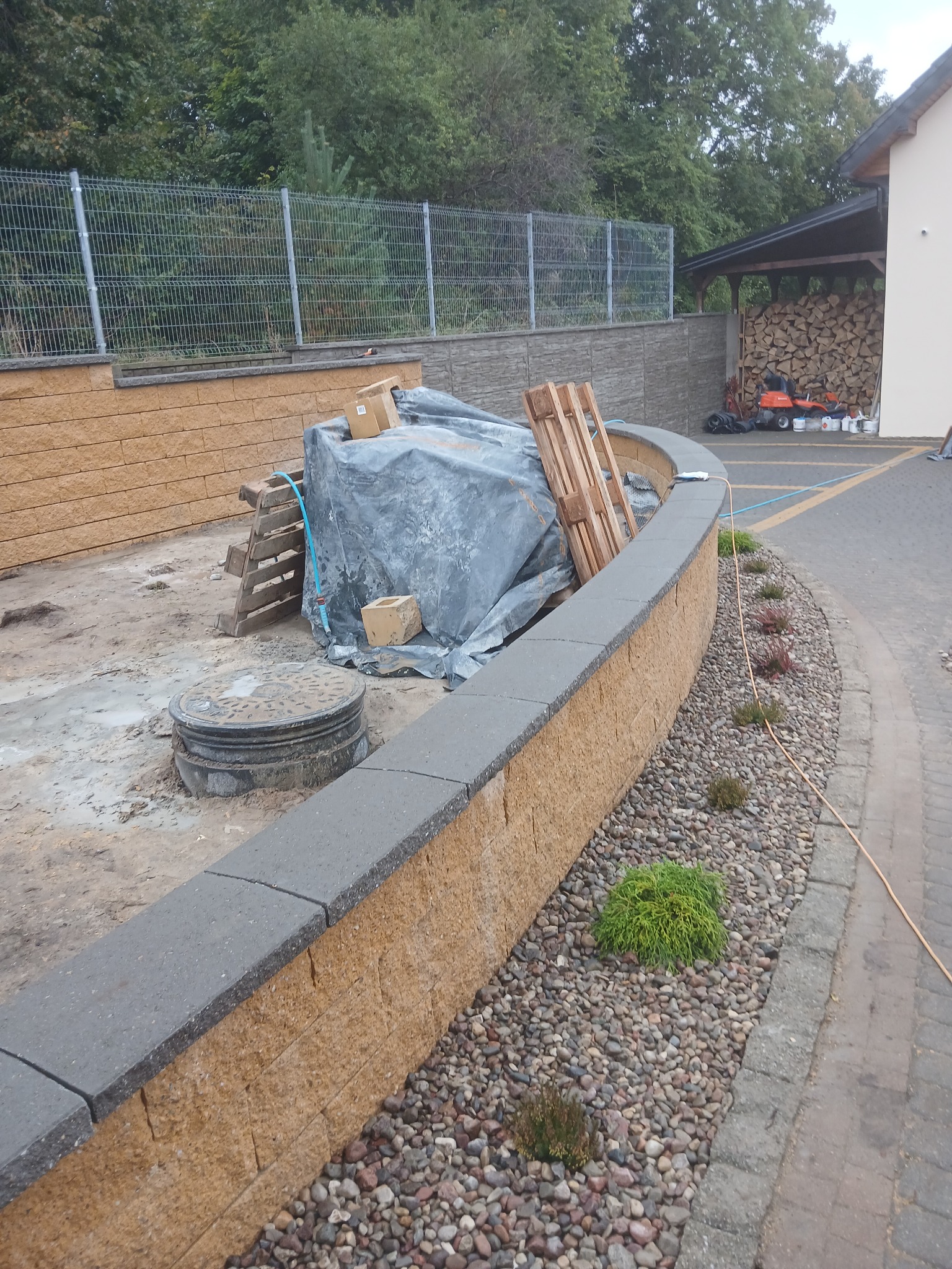 Mur oporowy z kamienia łupanego z szarą nakrywką, obok wysypane kamienie ozdobne i posadzone małe krzewy. W tle ogrodzenie panelowe i budynek gospodarczy.