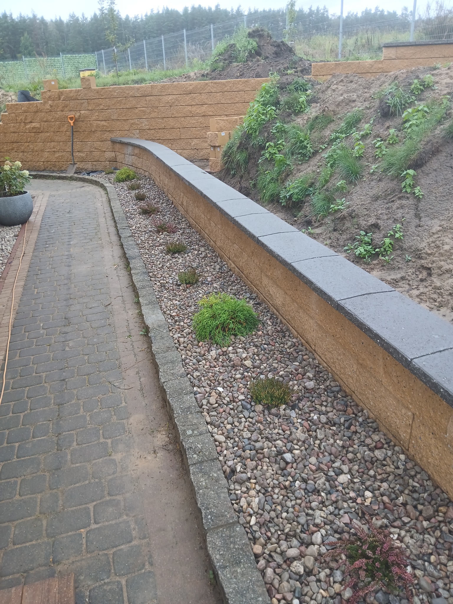 Wykończony murek oporowy z kamienia, oddzielający ścieżkę z kostki brukowej od skarpy. U podnóża murku ozdobne kamienie i rośliny. W tle siatka ogrodzeniowa.