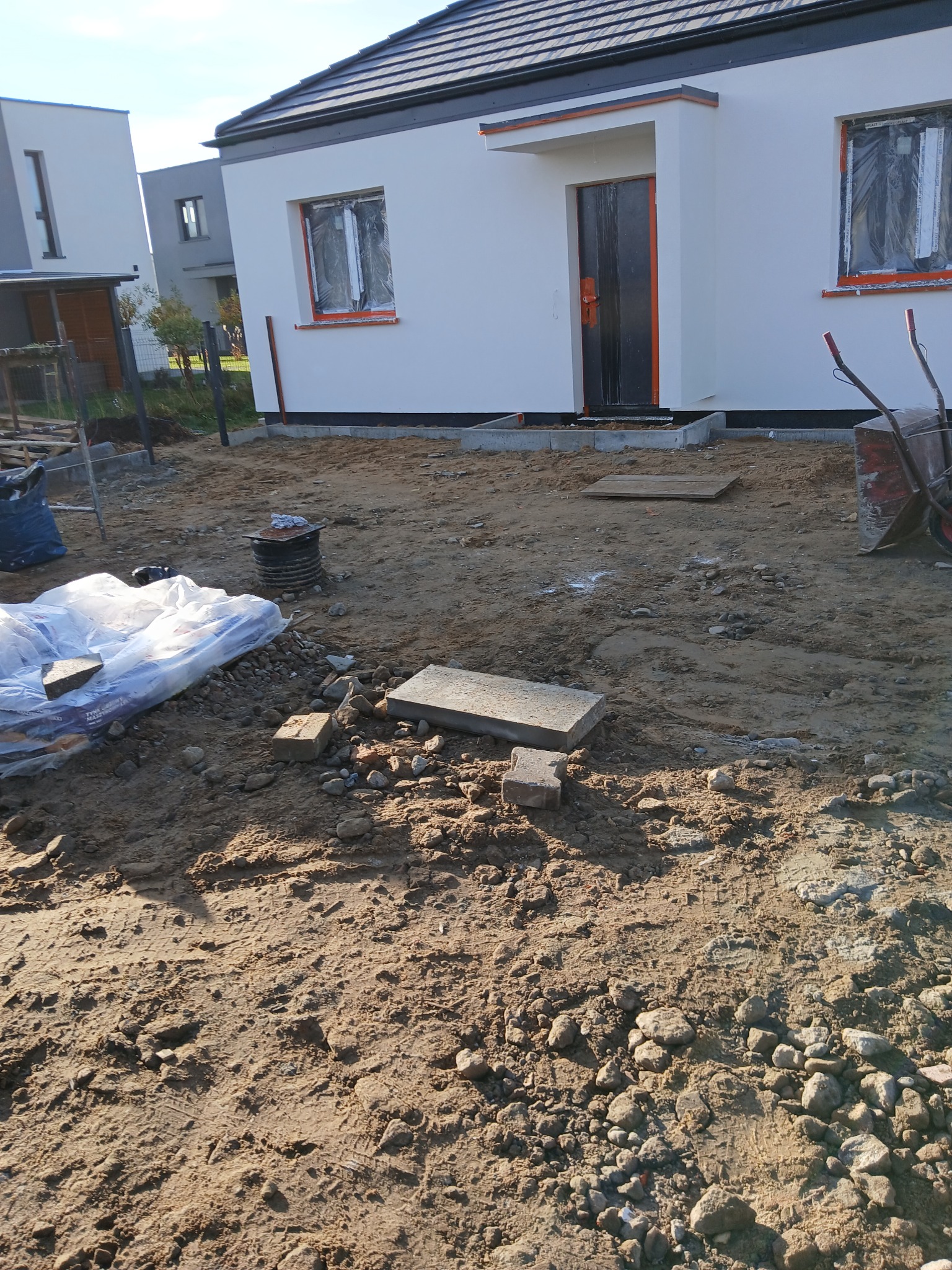 Nowy dom jednorodzinny w trakcie budowy, widoczne fundamenty i teren przygotowany pod kostkę brukową. Częściowo zakryte okna folią ochronną.