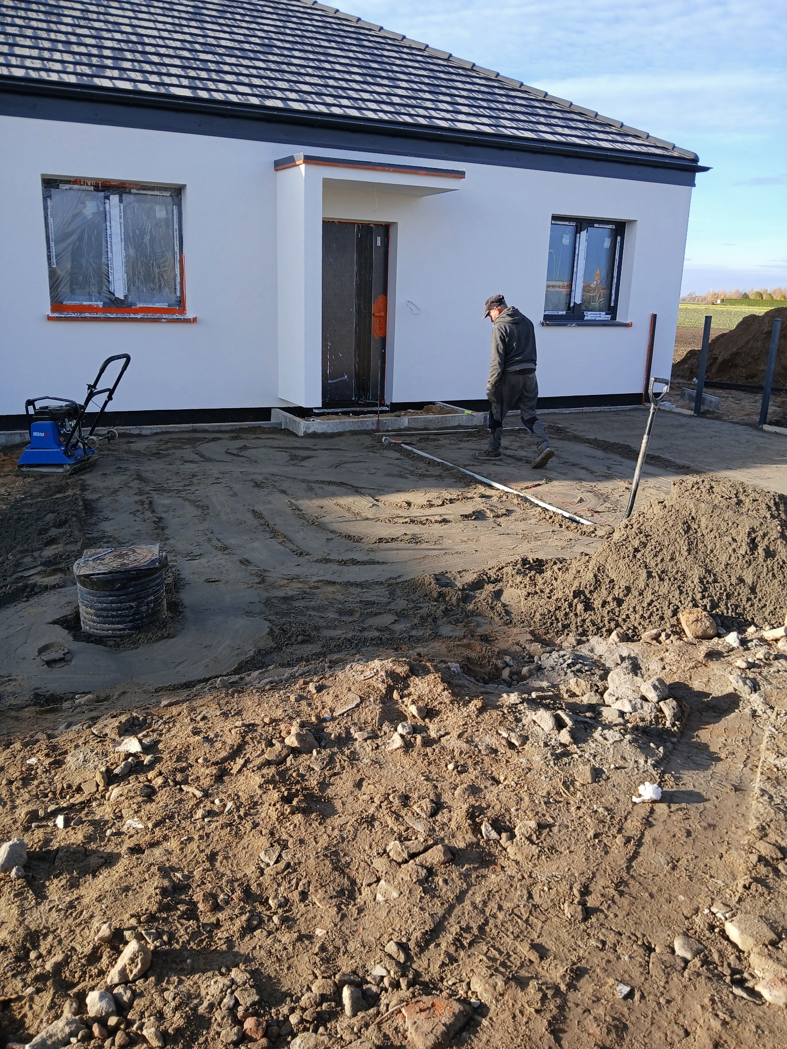Przygotowanie podłoża pod kostkę brukową przed domem jednorodzinnym. Widoczna ubijarka, piasek, oraz pracownik w trakcie wyrównywania terenu.