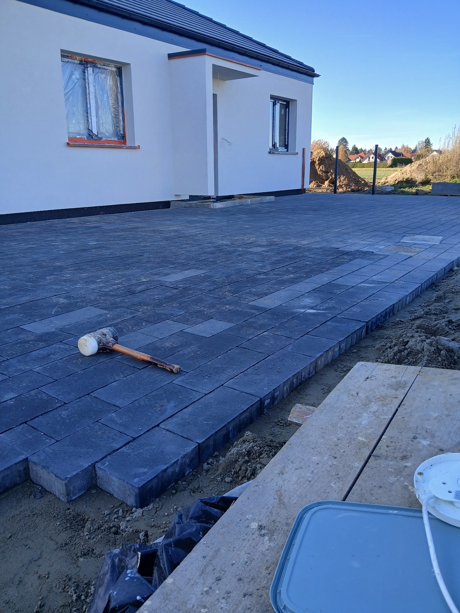 Ciemnoszara kostka brukowa w trakcie układania przed białym domem. Gumowy młotek leży na kostce. Widoczne narzędzia i materiały budowlane.