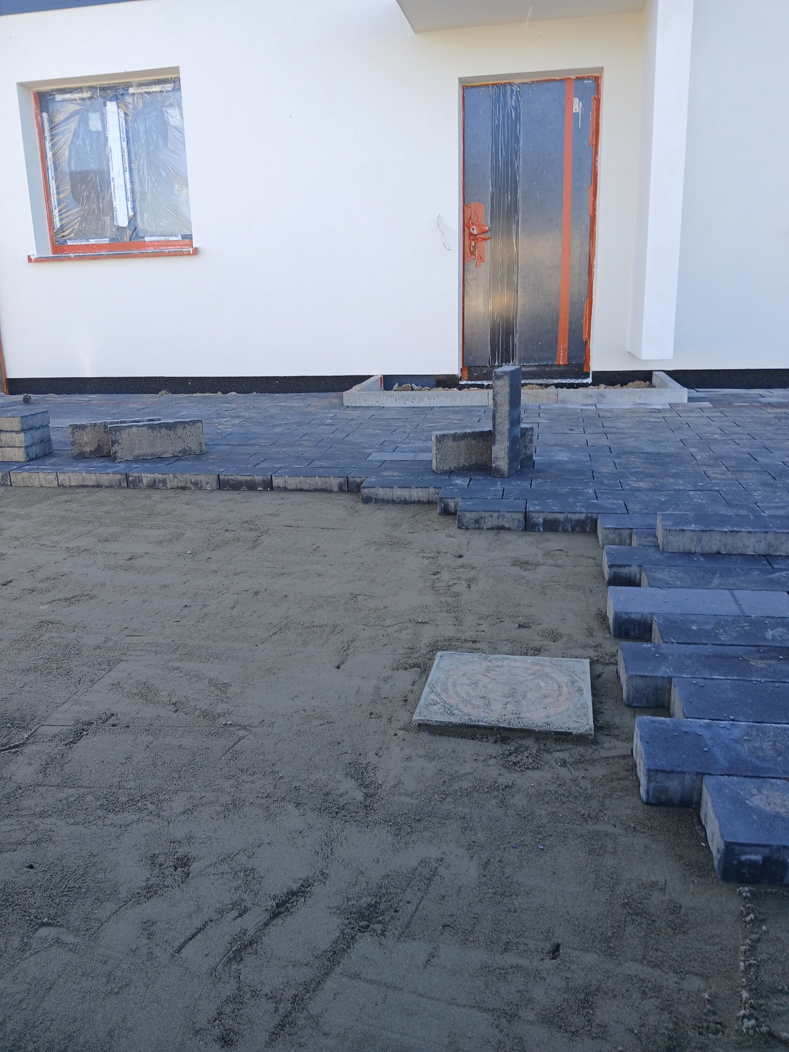 Częściowo ułożona, grafitowa kostka brukowa przed budynkiem z oknem i drzwiami zabezpieczonymi folią. Widoczny piasek i pojedyncze kostki. Ujęcie z dołu.