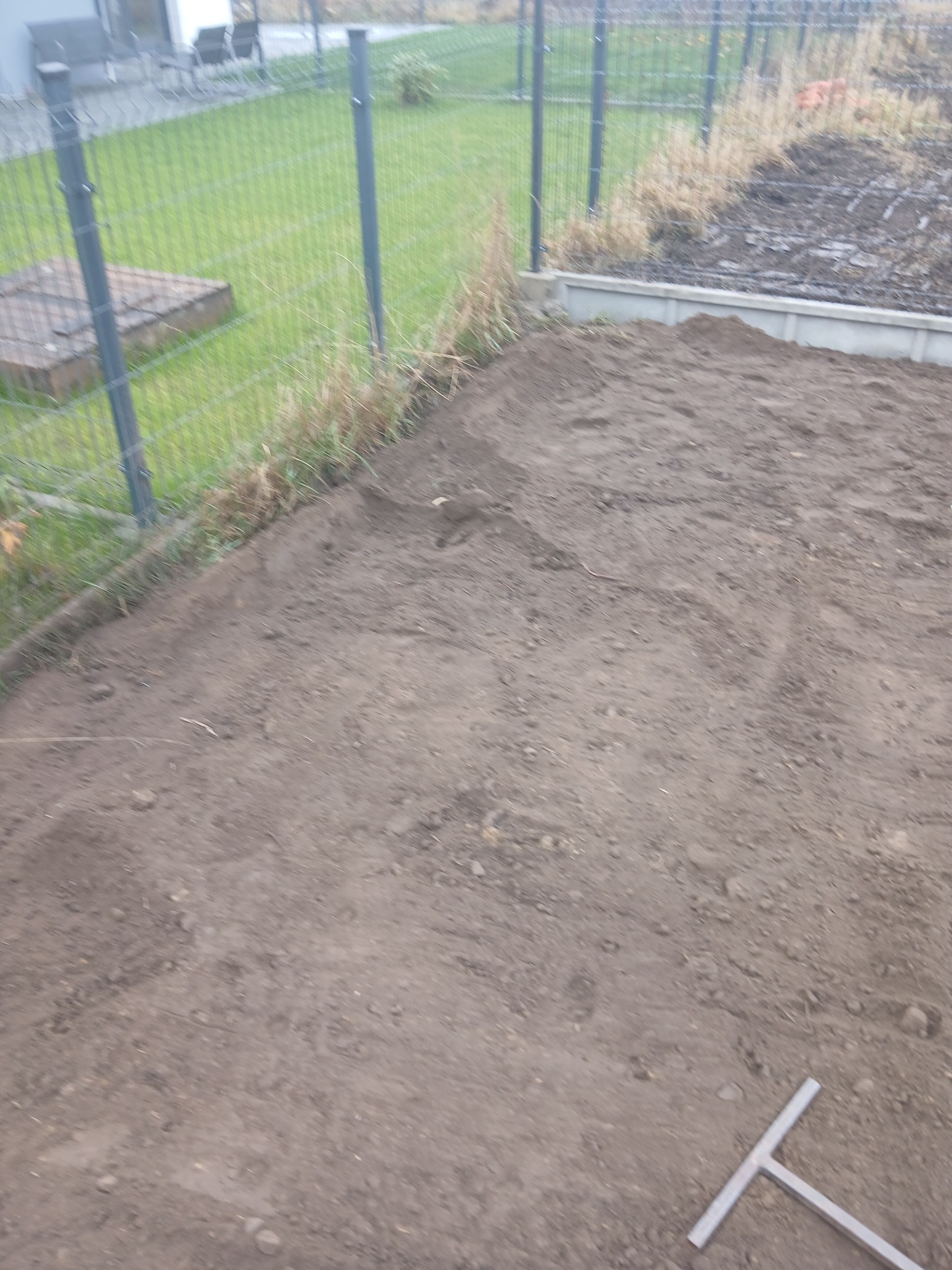 Przygotowany teren pod kostkę brukową z widocznym fragmentem ogrodzenia panelowego i narzędziem do wyrównywania gruntu w rogu kadru.