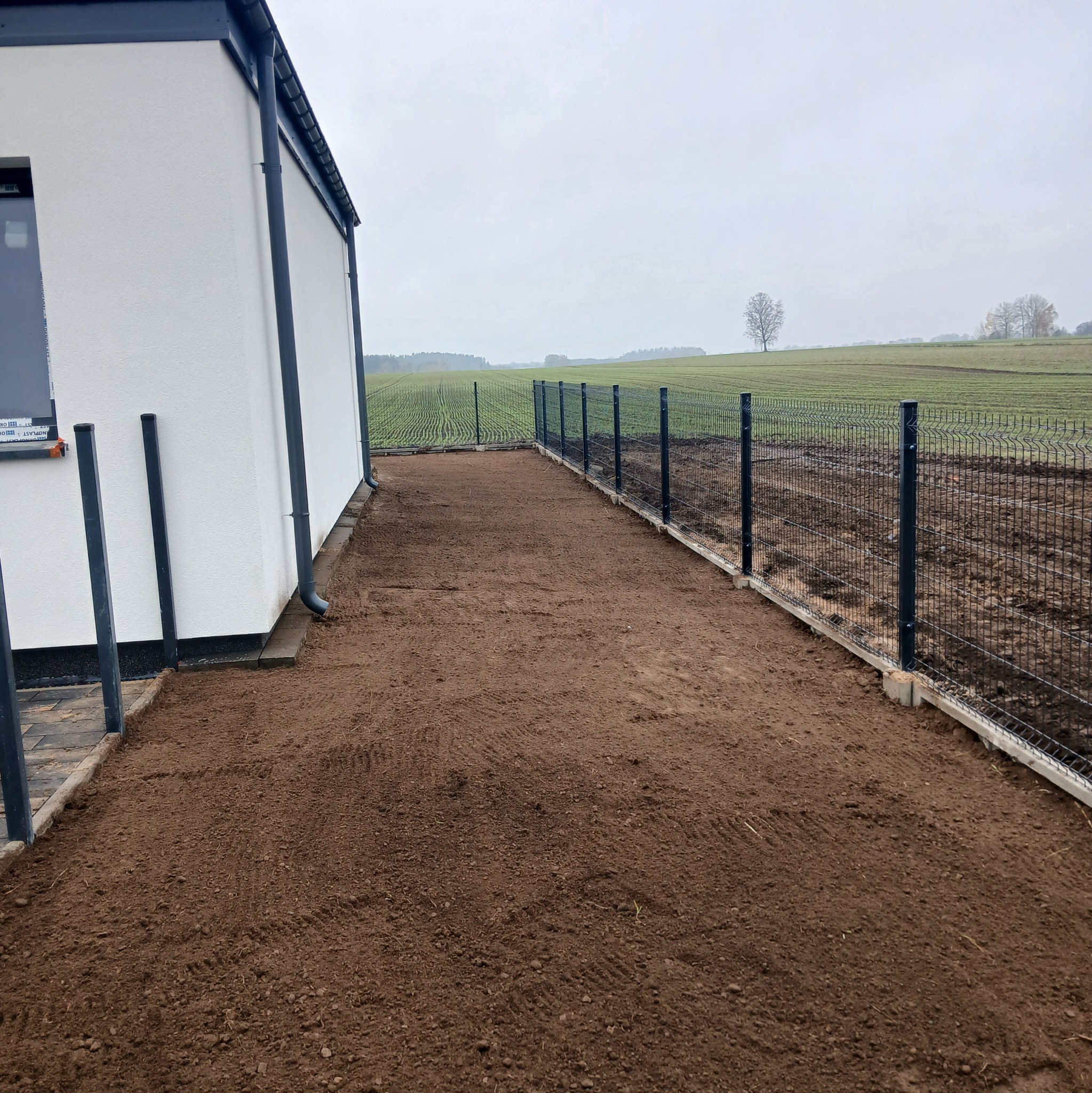 Nowoczesny dom z zamontowanym ogrodzeniem panelowym na podmurówce, przygotowany teren pod przyszły trawnik lub kostkę. Widok na pole w tle.