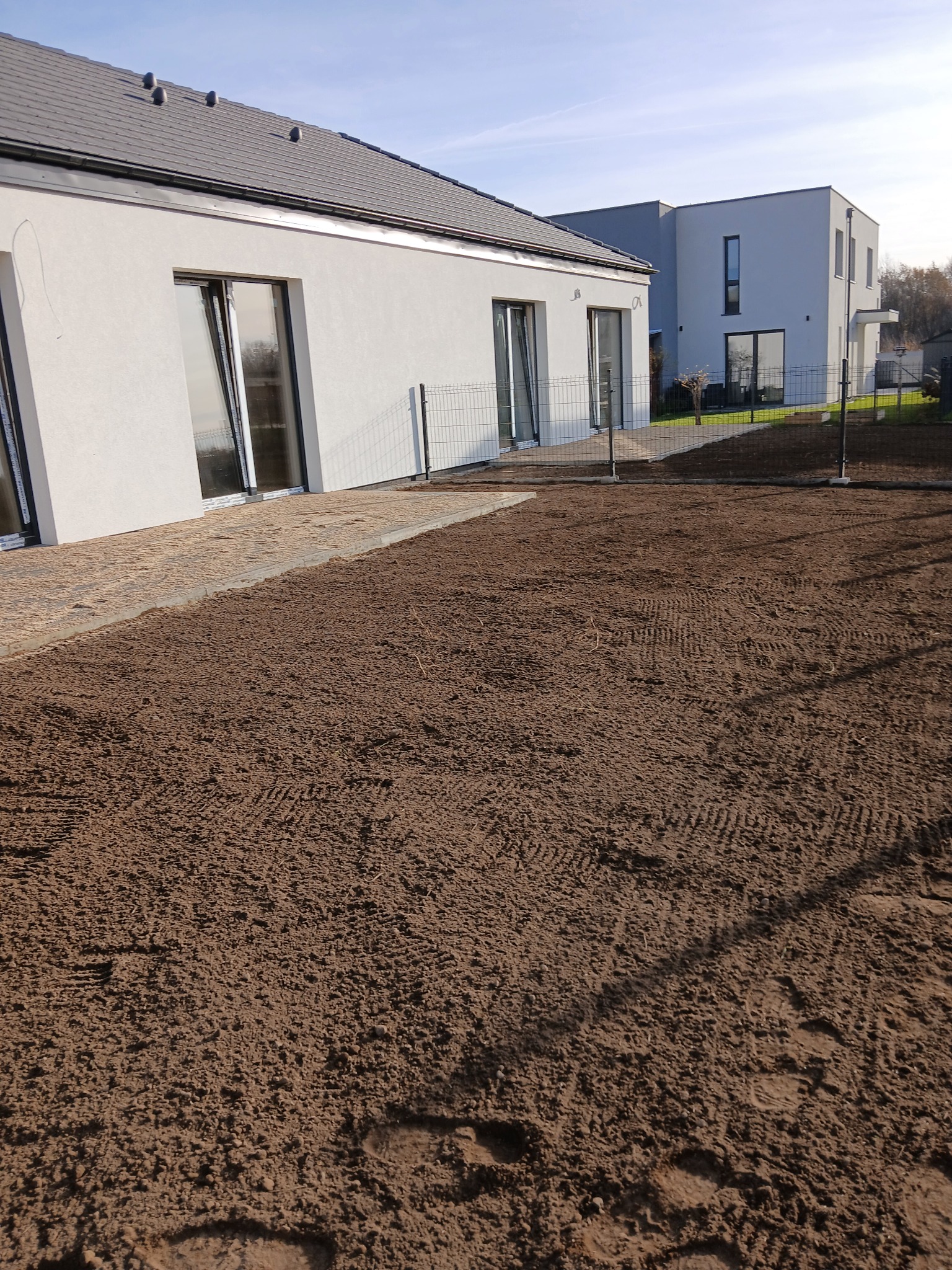 Przygotowany teren pod trawnik w Pobądzu. Widoczne dwa nowoczesne domy, ogrodzenie panelowe i wyrównana ziemia z śladami opon. Słoneczny dzień.