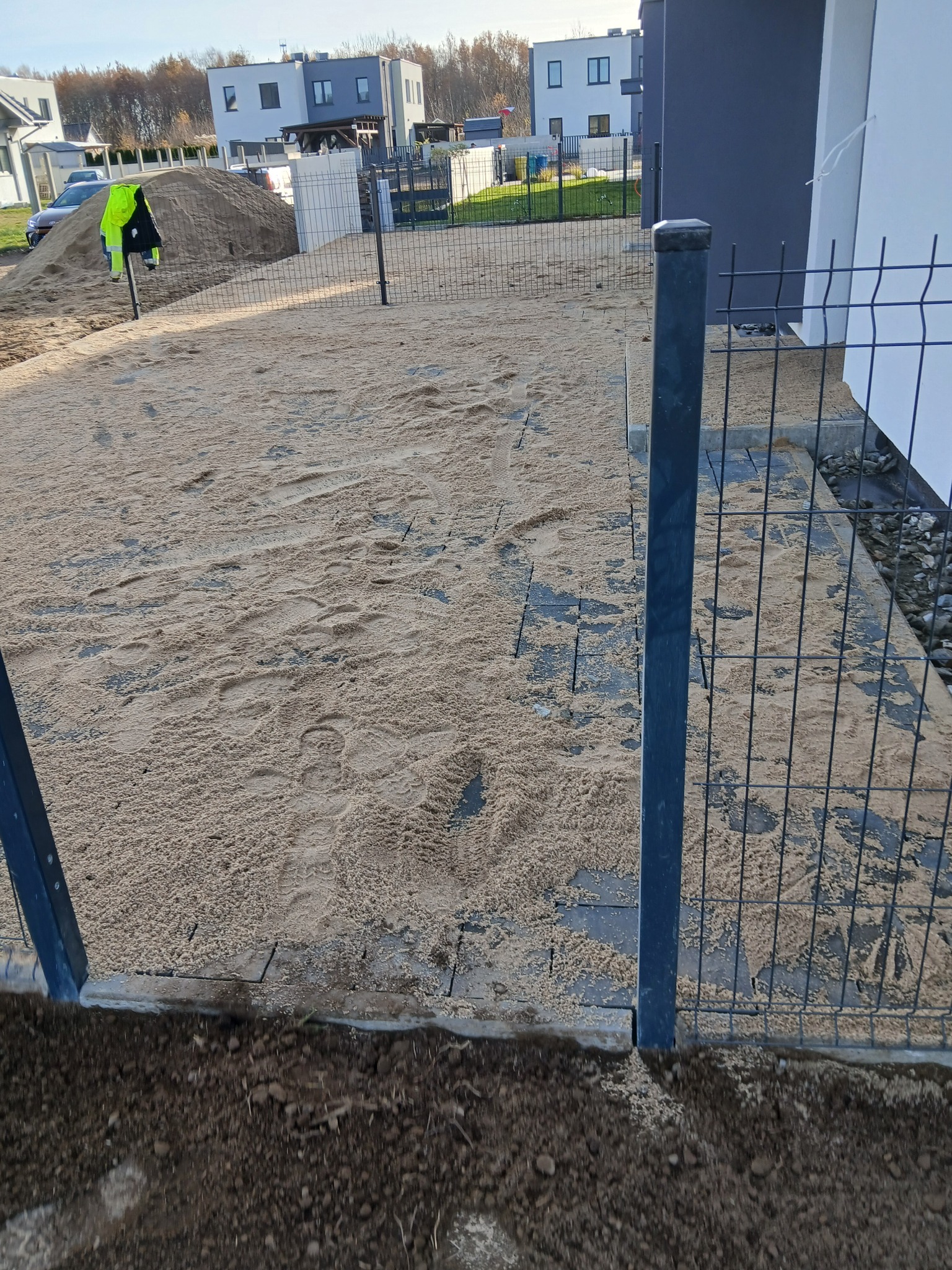 Montaż ogrodzenia panelowego na posesji z przygotowanym podłożem pod kostkę brukową. Widoczny piasek i częściowo ułożona kostka. W tle domy jednorodzinne.