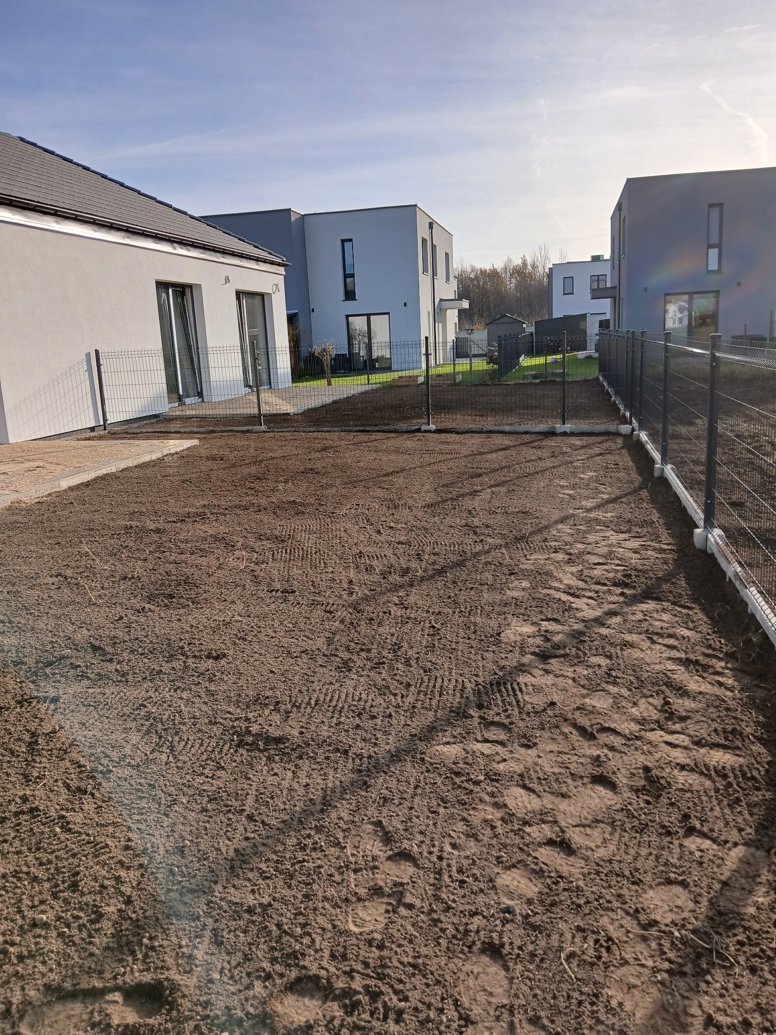 Przygotowany teren pod trawnik z częściowo zamontowanym ogrodzeniem panelowym na betonowej podmurówce, w tle nowoczesne domy jednorodzinne.