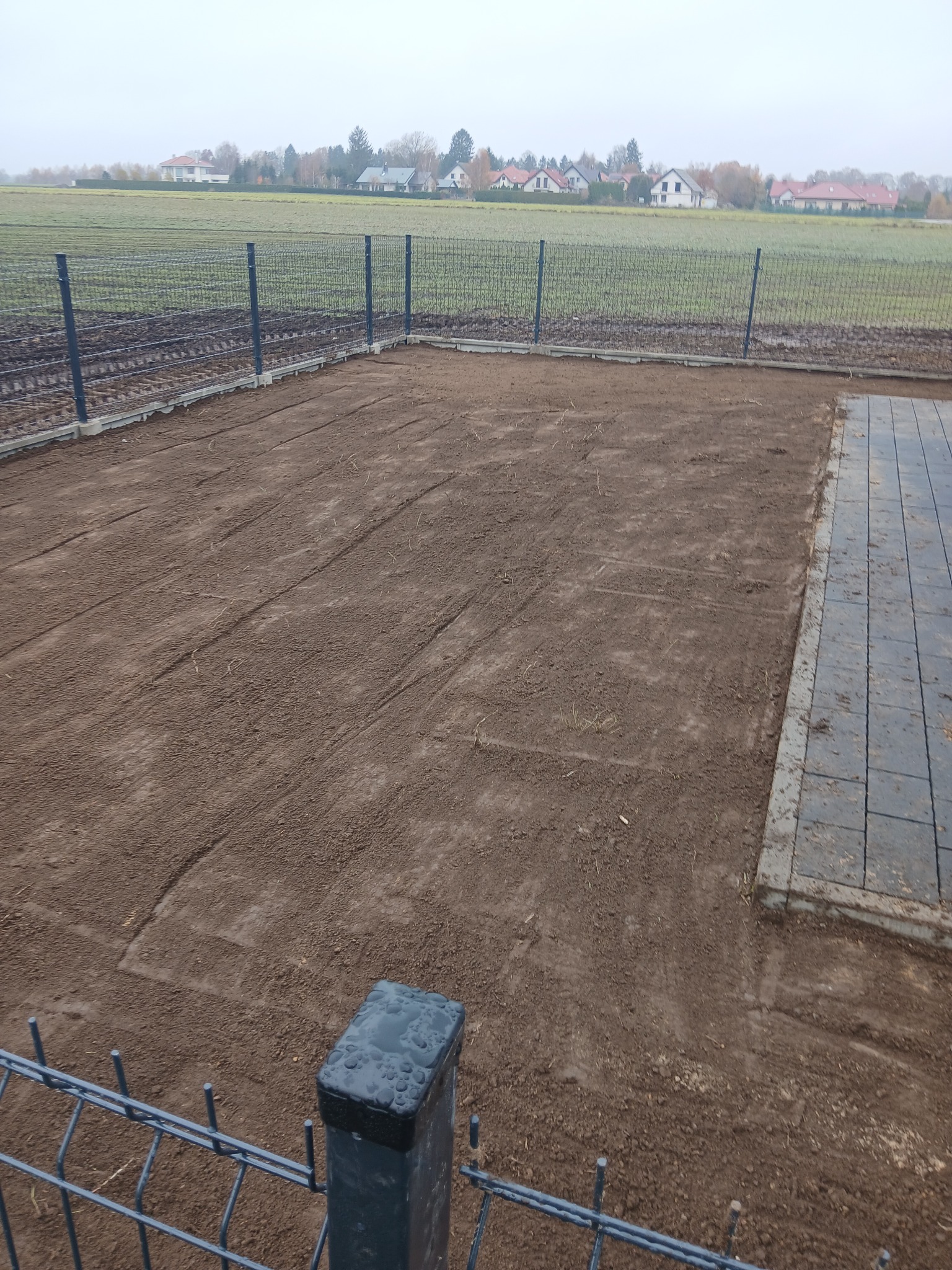 Przygotowany teren pod trawnik, obrzeża z kostki, ogrodzenie panelowe z metalową siatką na fundamencie. Widok na pole i zabudowania w tle, pochmurny dzień.
