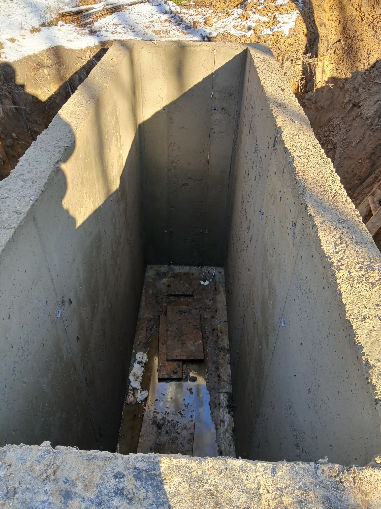 Betonowy wykop fundamentowy z widocznymi śladami wody i ziemi na dnie, oświetlony z jednej strony, z fragmentem śniegu w tle. Surowy, industrialny charakter.